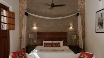 A bedroom with a bed, two chairs, a small table, a ceiling fan, and a wall-mounted light fixture.