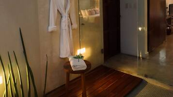 A hotel room with a wooden floor, a white robe hanging on the wall, a small table with a towel, and a large mirror reflecting the room.