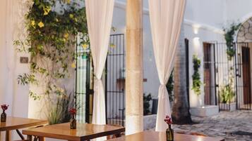 Un área de comedor al aire libre con mesas y sillas de madera, cortinas blancas y un motivo solar decorativo en la ventana.