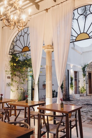 An outdoor dining area with wooden tables and chairs, white curtains, and a decorative sun motif on the window. - Casa de Las Palomas by Palomas Hotels -  Adults only (Mérida)