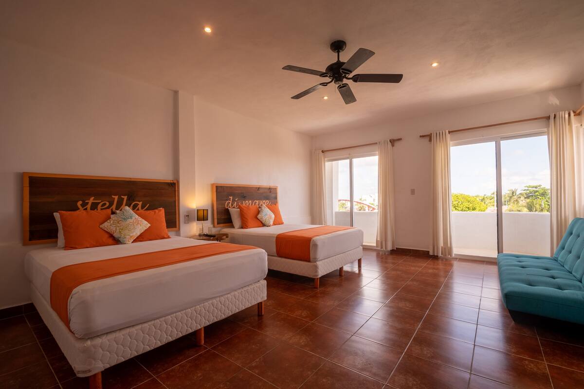 Habitación de hotel con dos camas, ventilador de techo, balcón con cortinas y un sofá color turquesa.