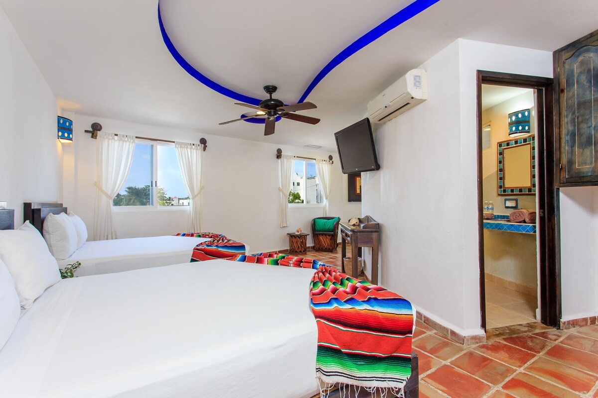 Habitación de hotel con dos camas, ventilador de techo, televisor y vista al exterior.