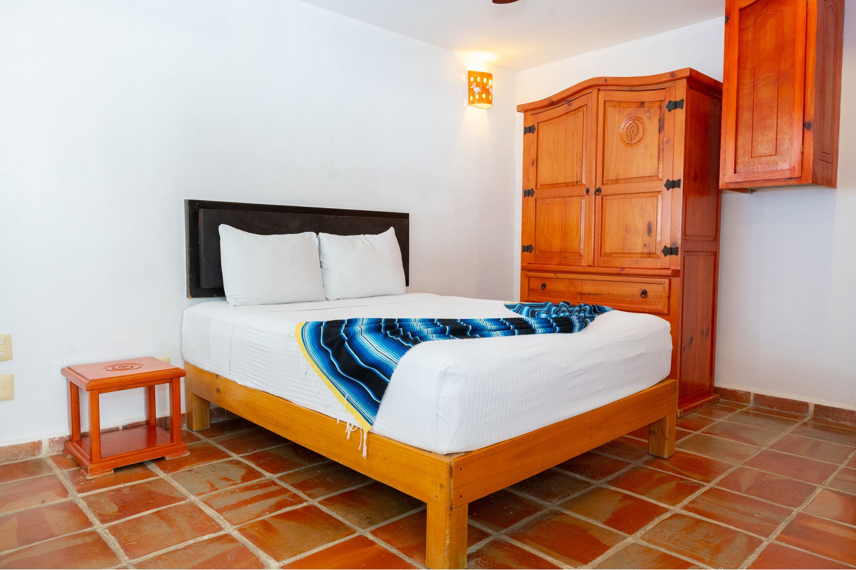 A bed with a white mattress and a blue and white striped blanket, a wooden headboard, a small wooden nightstand, and a wooden wardrobe with a mirror.