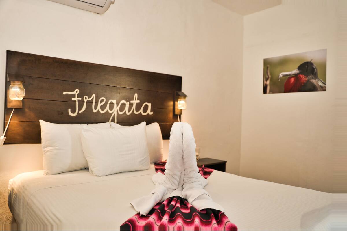 Habitación de hotel con una cama, almohadas blancas, cabecero con el nombre 'Fregata', manta roja y negra, y un cuadro en la pared.