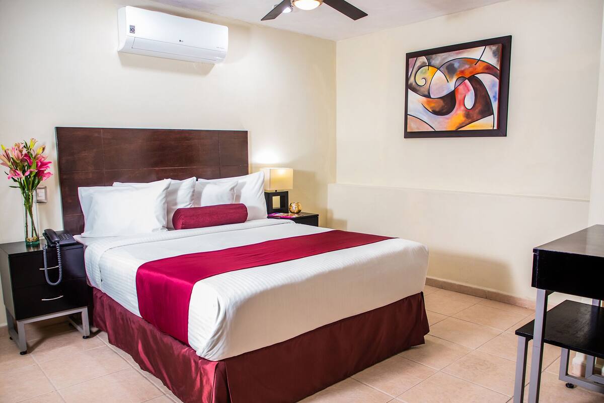 A hotel room with a bed, a nightstand, a desk, an air conditioner, and a painting on the wall.