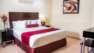Una habitación de hotel con cama, mesita de noche, escritorio, aire acondicionado y un cuadro en la pared.
