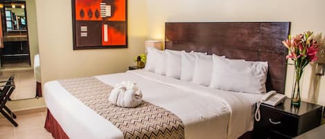 A hotel room with a large bed, a ceiling fan, a wall-mounted air conditioner, a painting, a bedside table with a phone, and a vase with flowers.