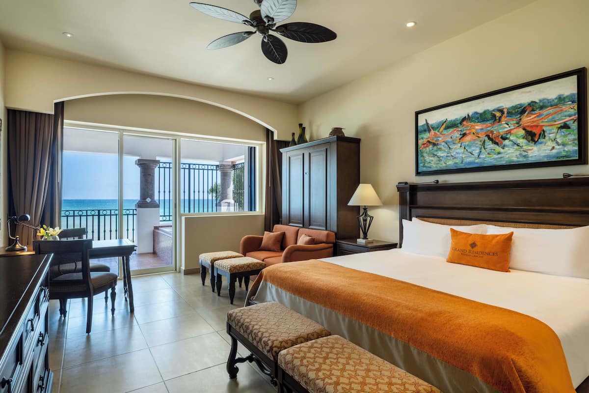 A hotel room with a large bed, a desk, a chair, a sofa, and a balcony with a view of the ocean.
