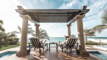 A covered outdoor seating area with a pool, palm trees, and a view of the ocean.