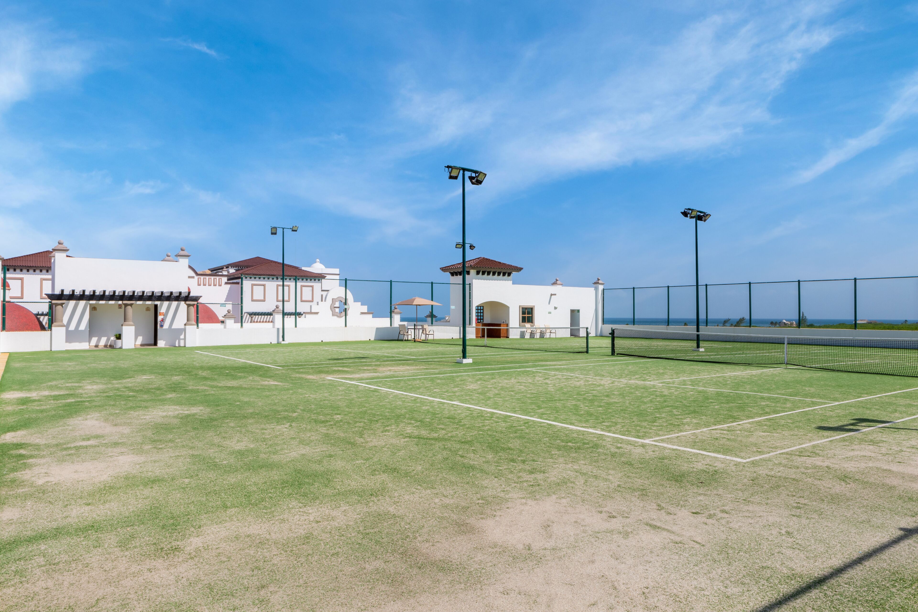A tennis court with a net, surrounded by a fence, and a building with a red roof in the background.