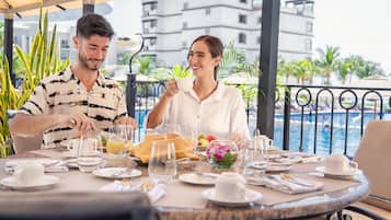 Mesa de comedor lista para dos personas, con vista a una piscina y edificios al fondo.