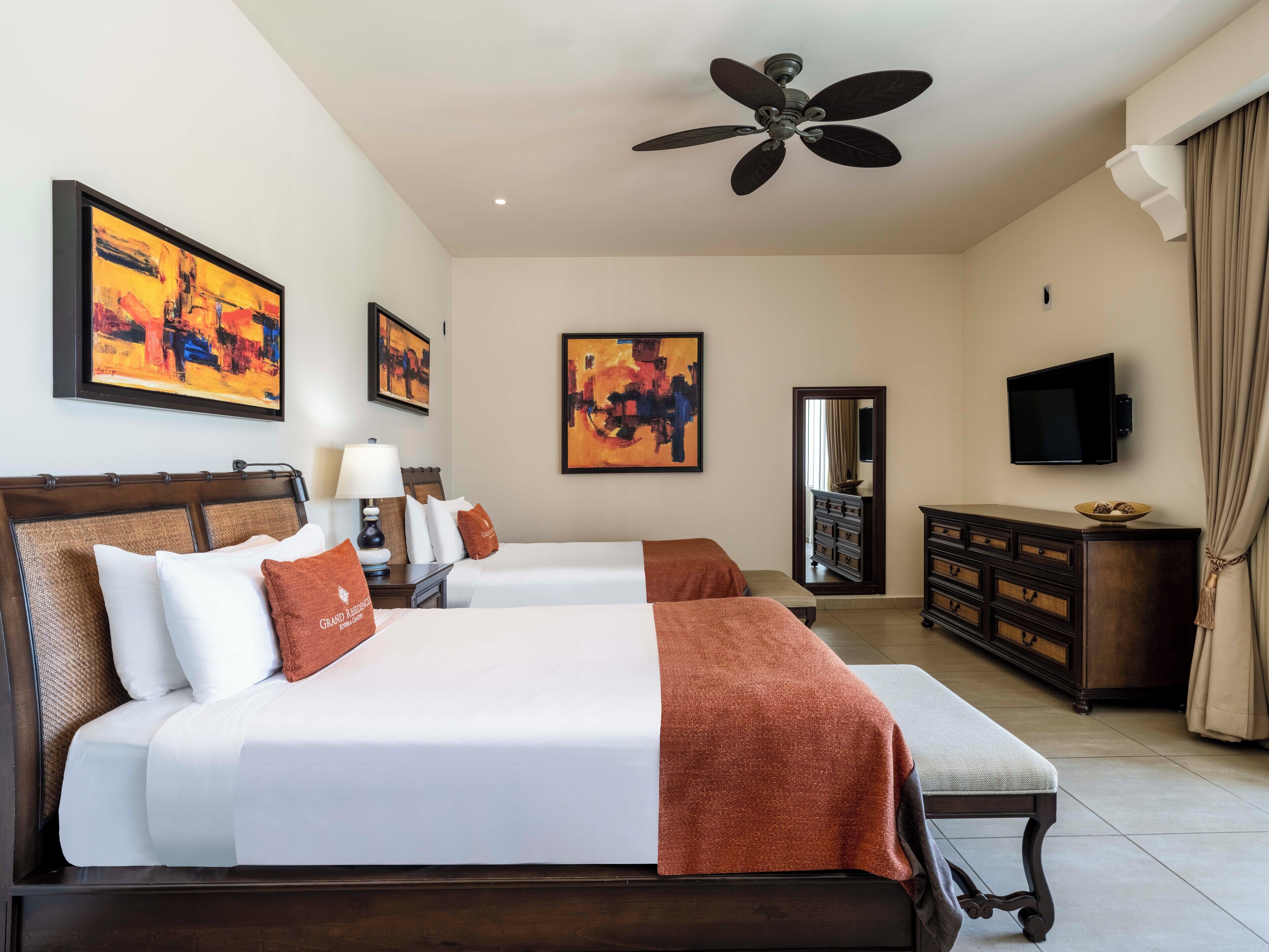 A hotel room with two beds, a ceiling fan, artwork on the walls, a television, and a dresser.