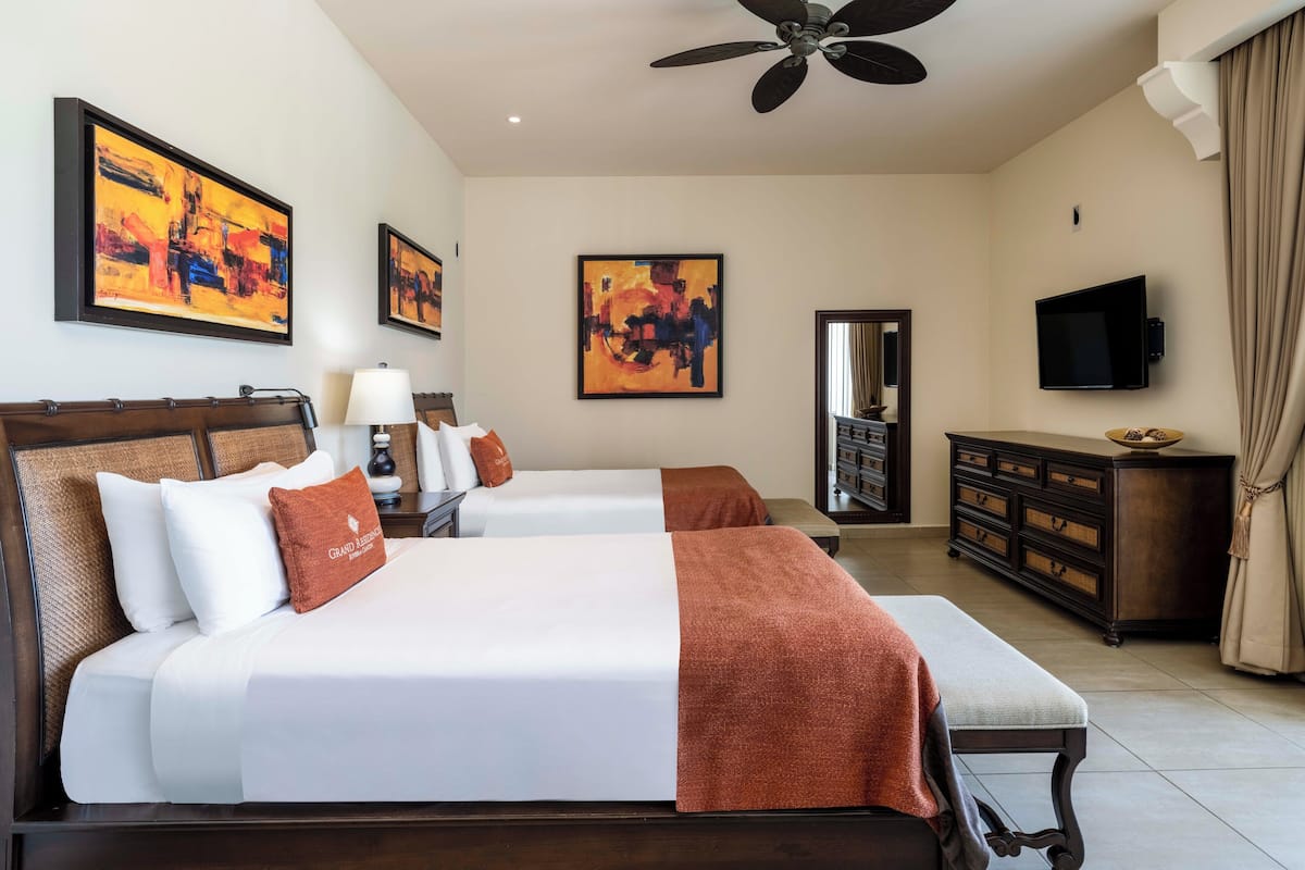 A hotel room with two beds, a ceiling fan, artwork on the walls, a television, and a dresser.