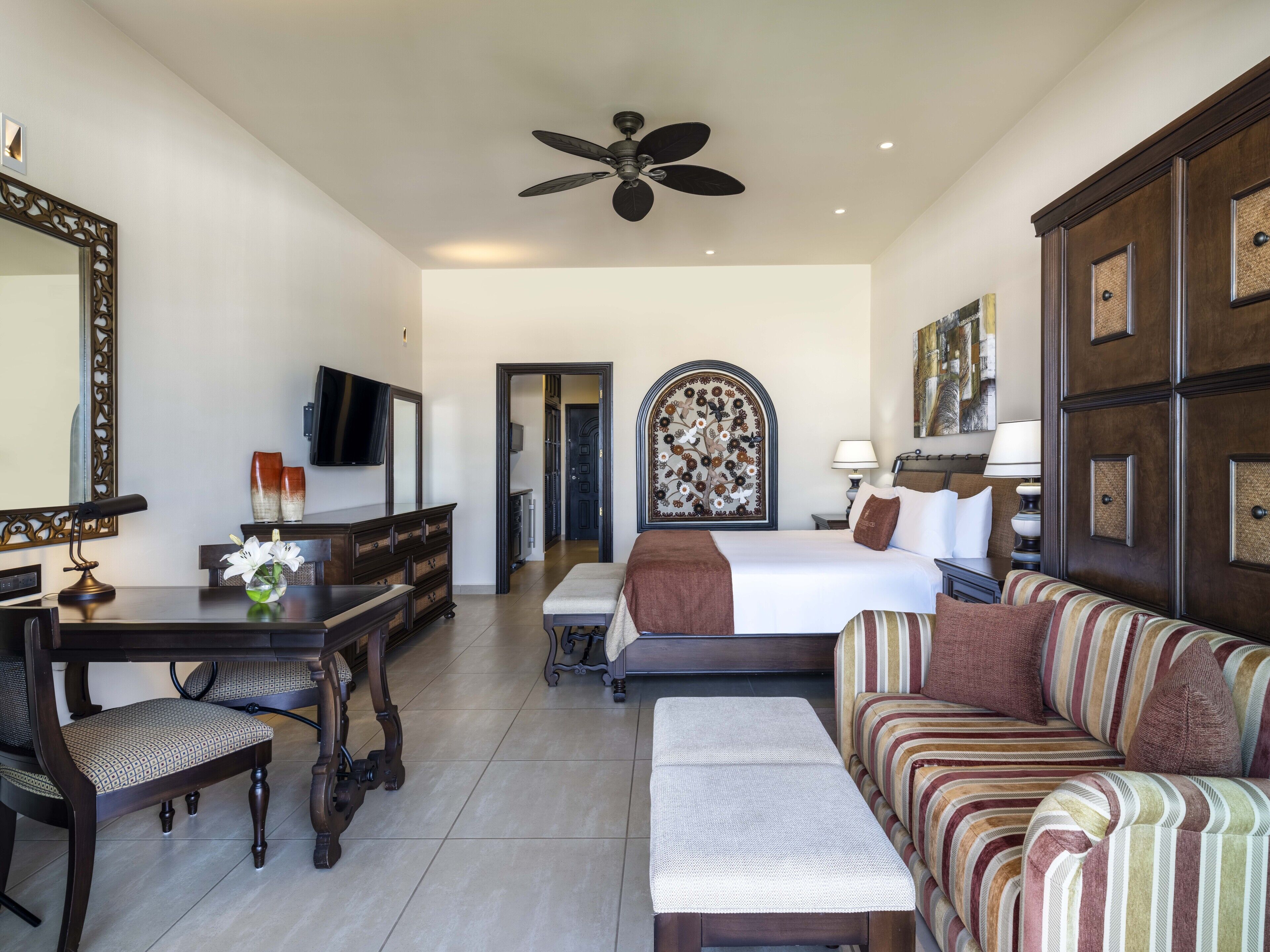 A hotel room with a bed, a sofa, a desk with a chair, a television, and a ceiling fan.