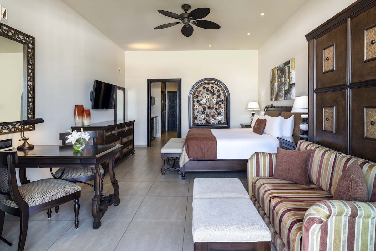 A hotel room with a bed, a sofa, a desk with a chair, a television, and a ceiling fan.