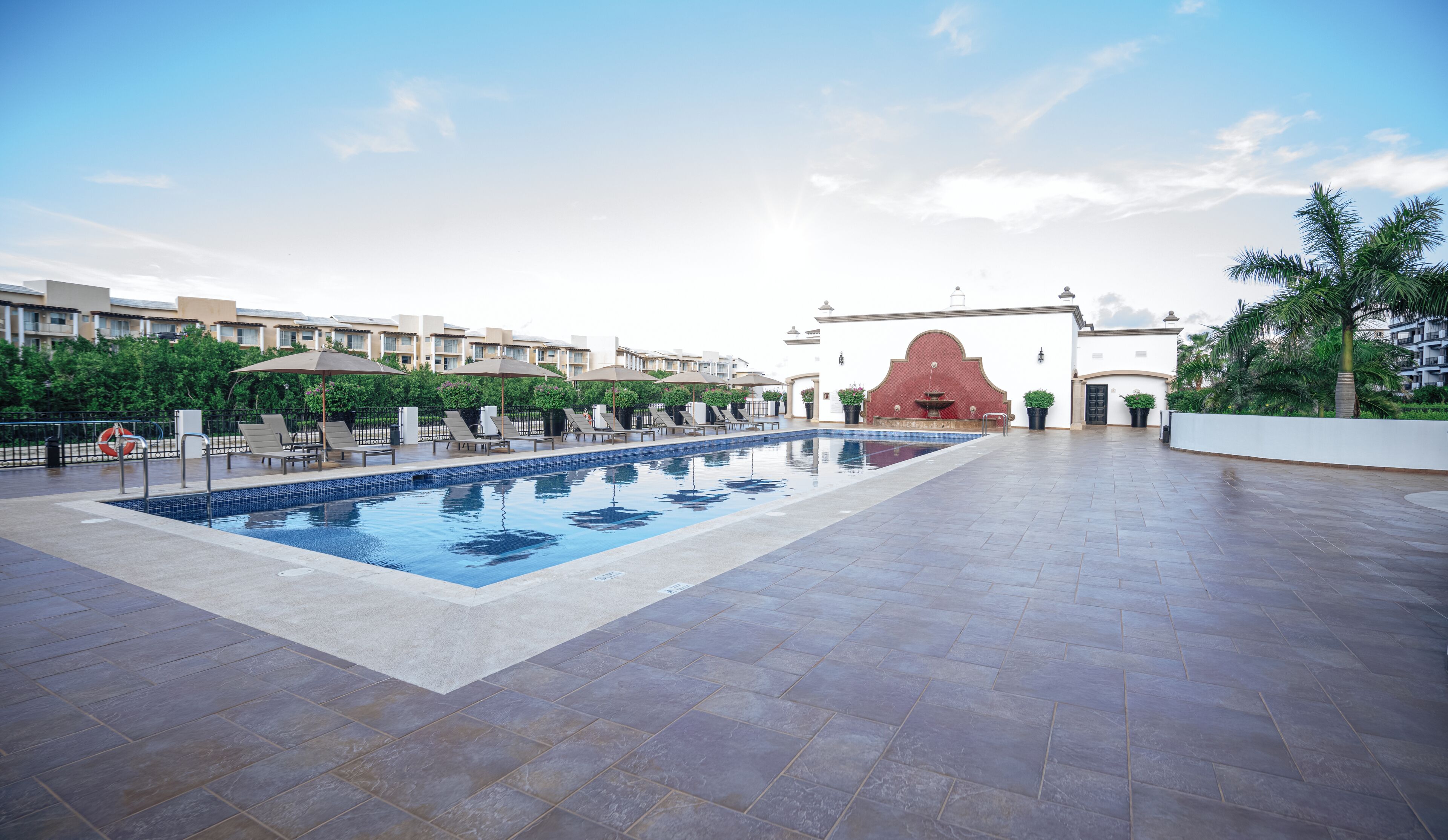 Piscina con sillas de descanso y sombrillas, rodeada por un patio empedrado y un edificio de fachada roja.