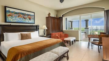 A hotel room with a large bed, a desk, a chair, a bench, a ceiling fan, a painting, a cabinet, and a balcony with a view.