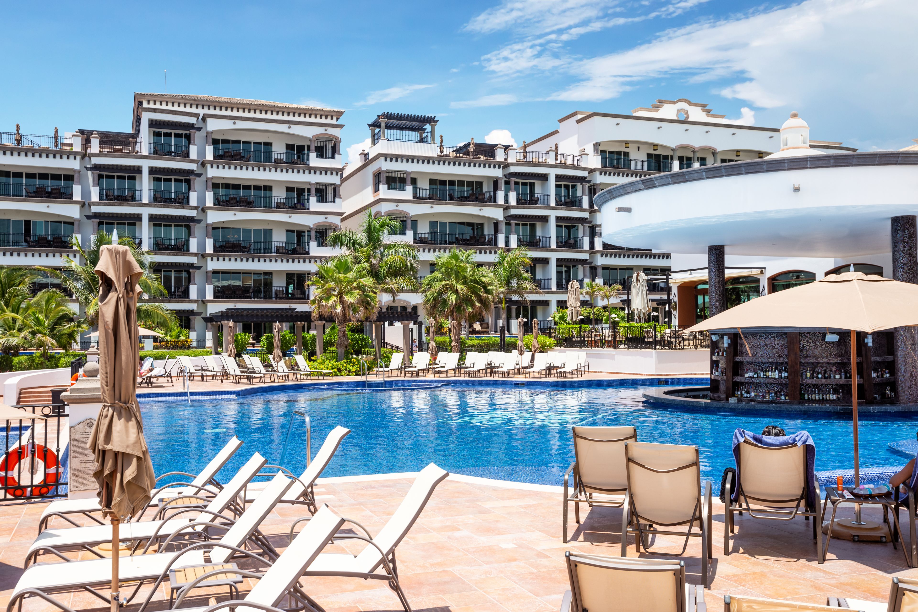 Zona de piscina de hotel con múltiples sillones y sombrillas, rodeada por un edificio grande con balcones.
