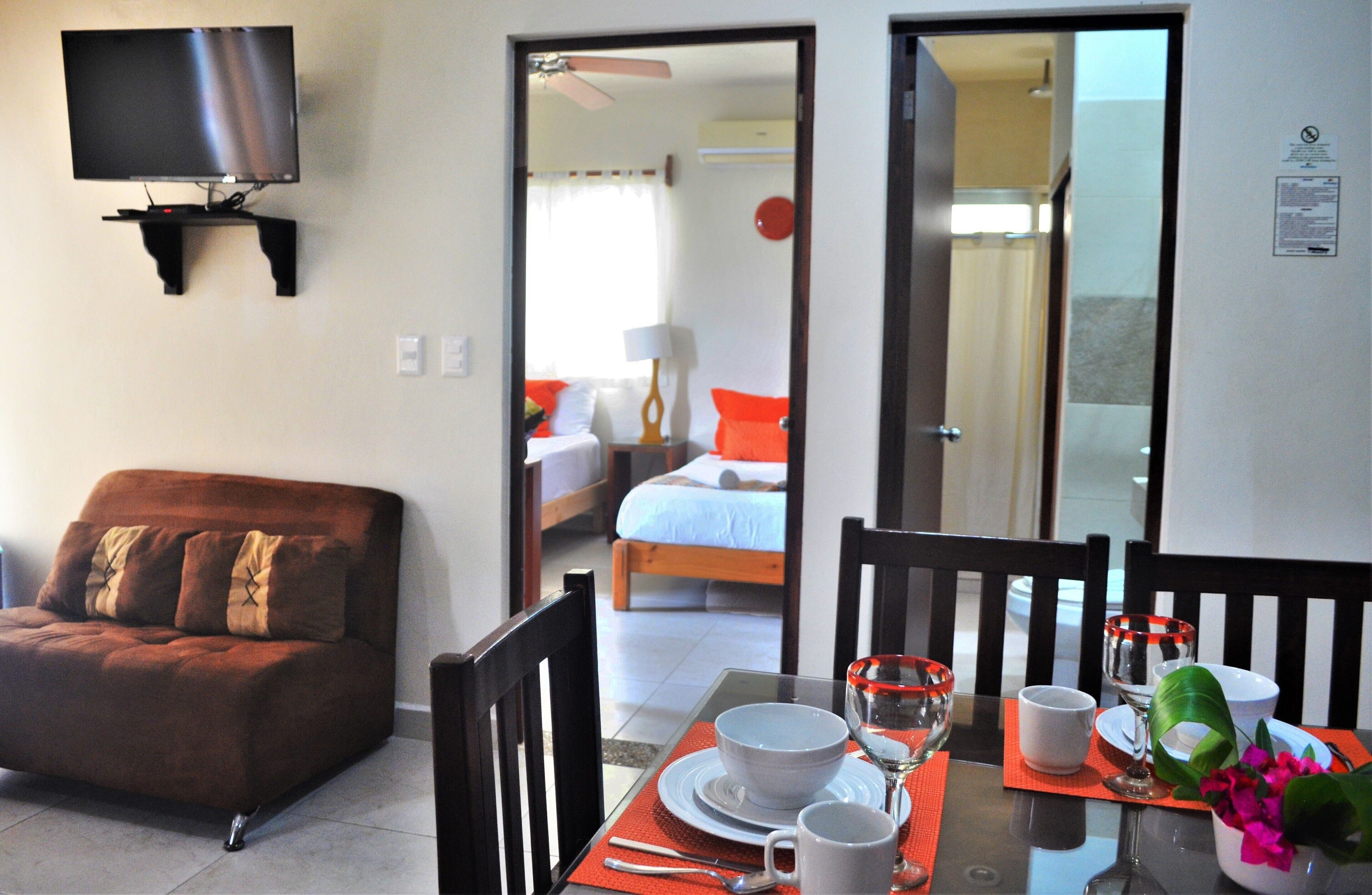 A hotel room with a TV mounted on the wall, a dining table set with plates, glasses, and a bowl of flowers.