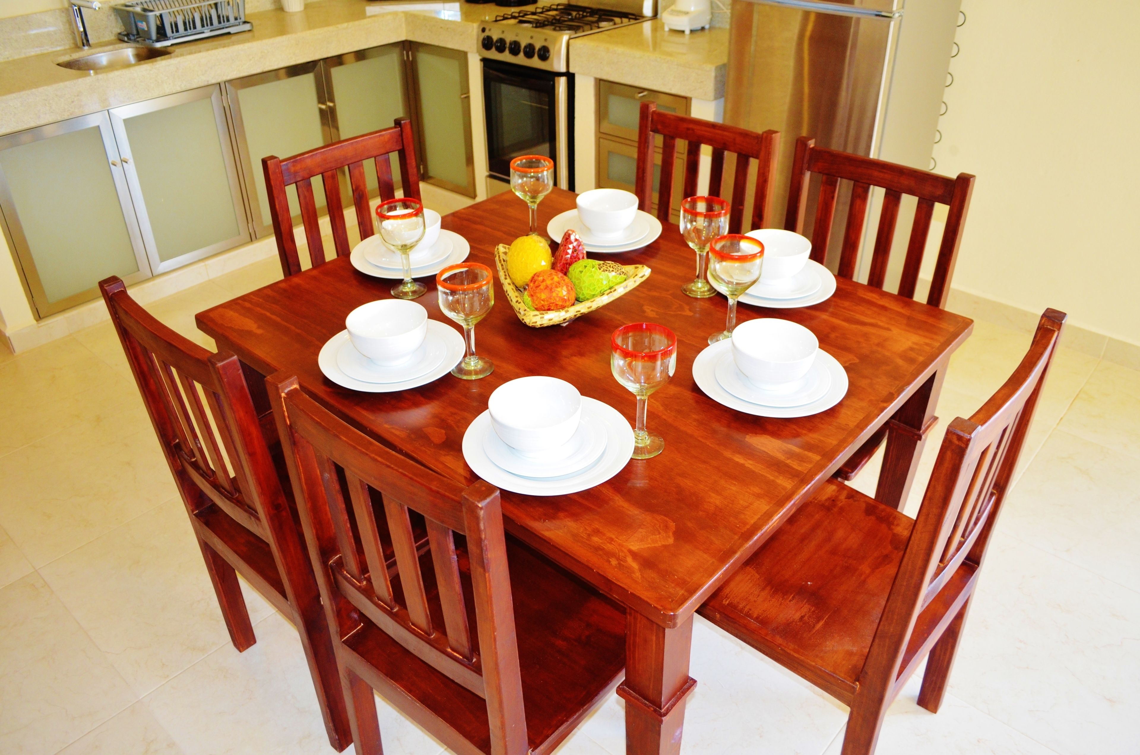 Una mesa de comedor de madera dispuesta con platos, cuencos y vasos blancos, rodeada de sillas de madera.