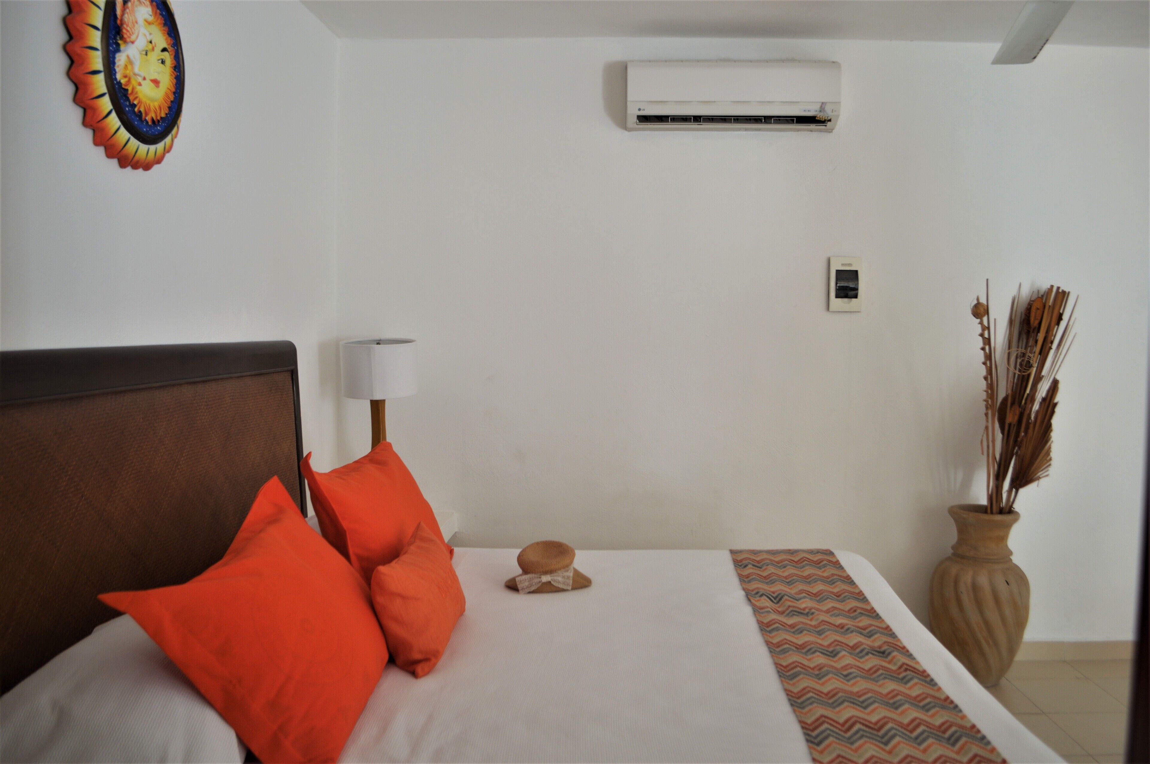 Una camera d'albergo con un letto, cuscini, una lampada da comodino, un condizionatore d'aria, una decorazione a parete, un vaso con piante essiccate e un tappeto a motivi.