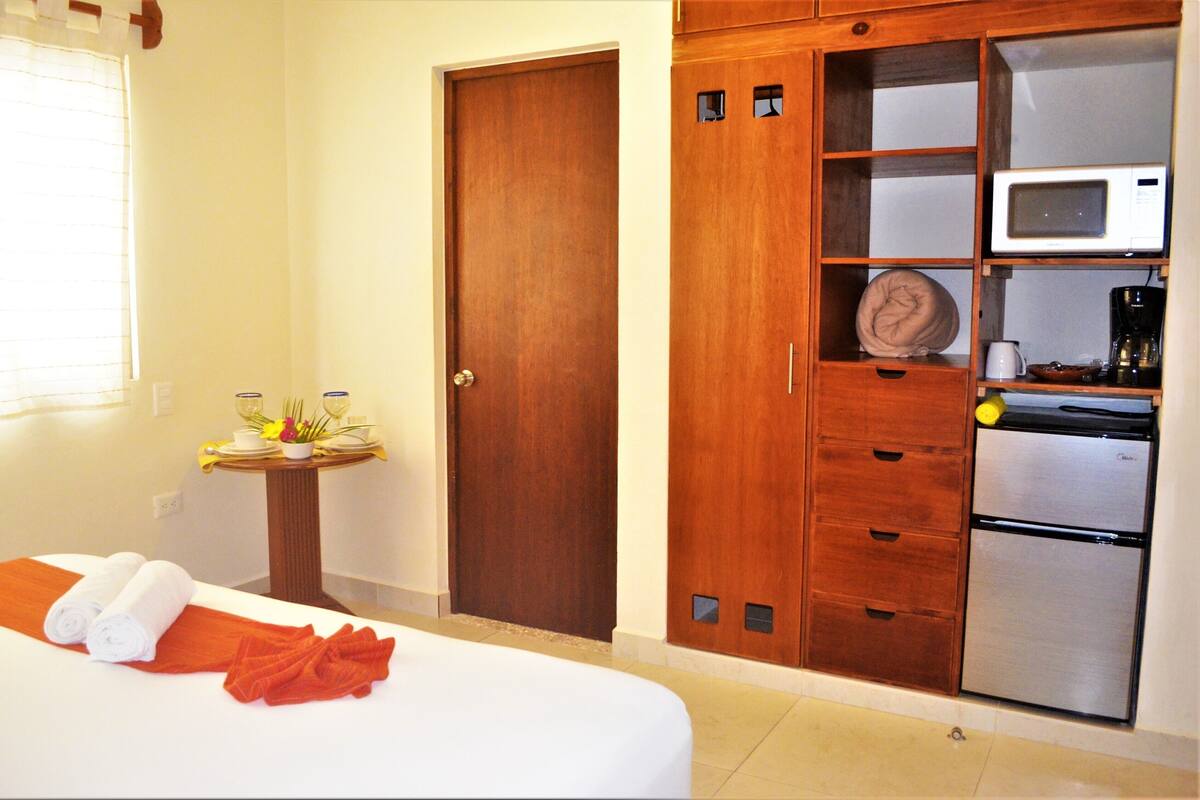 Habitación de hotel con cama, armario de madera, microondas, cafetera y una pequeña mesa con un jarrón y flores.