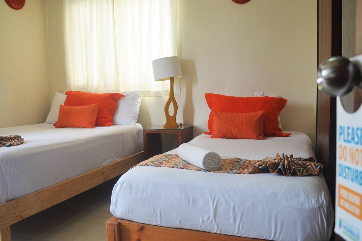 Dos camas individuales con ropa de cama blanca y naranja, una mesita de noche de madera con una lámpara, y un cartel en la puerta.