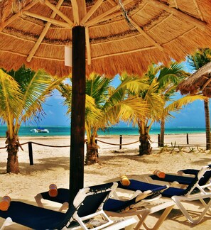 A beach scene with thatched umbrellas, palm trees, and lounge chairs. - Hotel Arrecifes Suites (Puerto Morelos)
