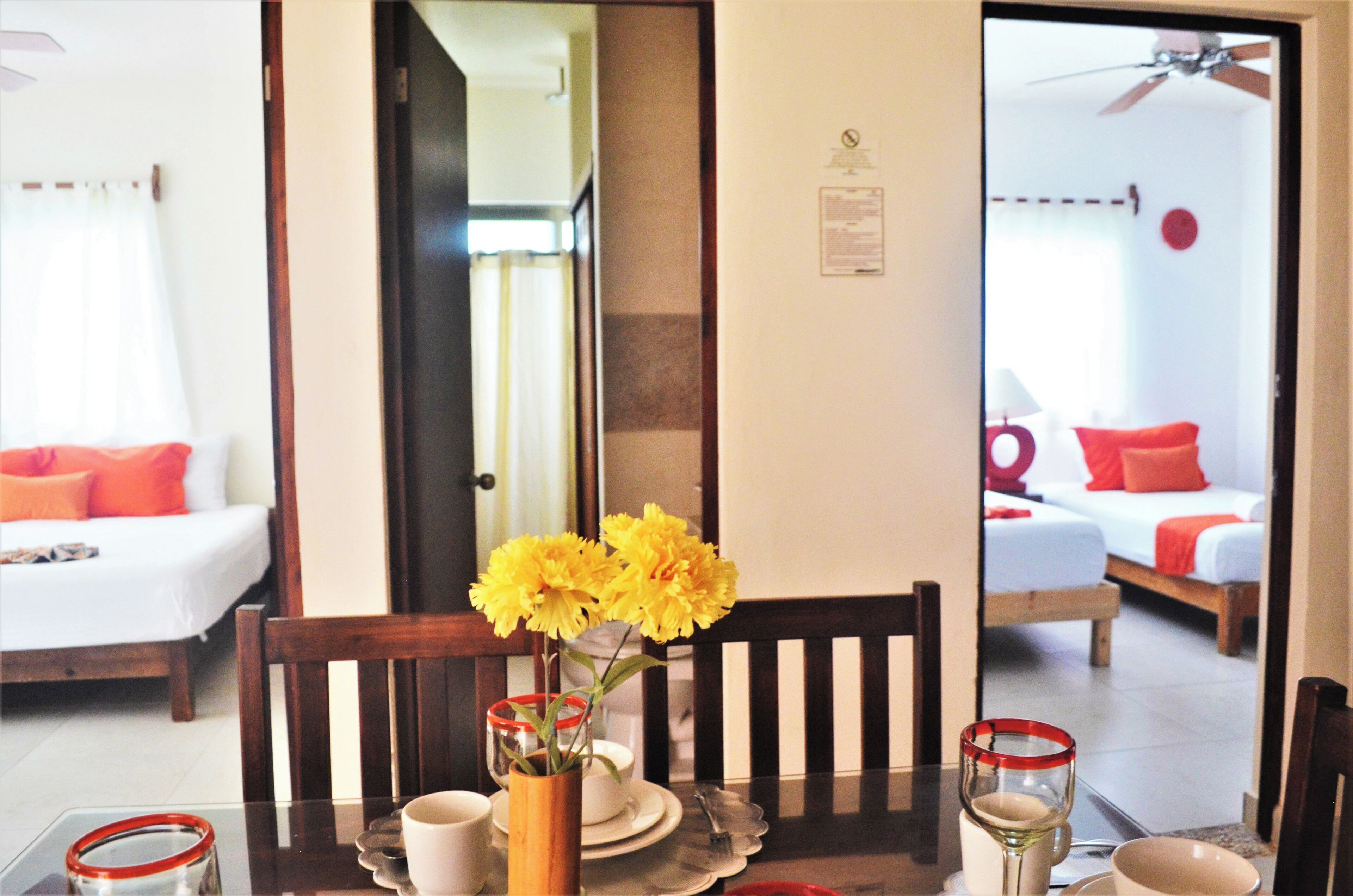 Un coin repas avec une table dressée pour deux personnes, un vase rempli de fleurs jaunes et une vue sur une chambre à coucher avec un lit et un oreiller rouge.