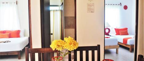 Un coin repas avec une table dressée pour deux personnes, un vase rempli de fleurs jaunes et une vue sur une chambre à coucher avec un lit et un oreiller rouge.