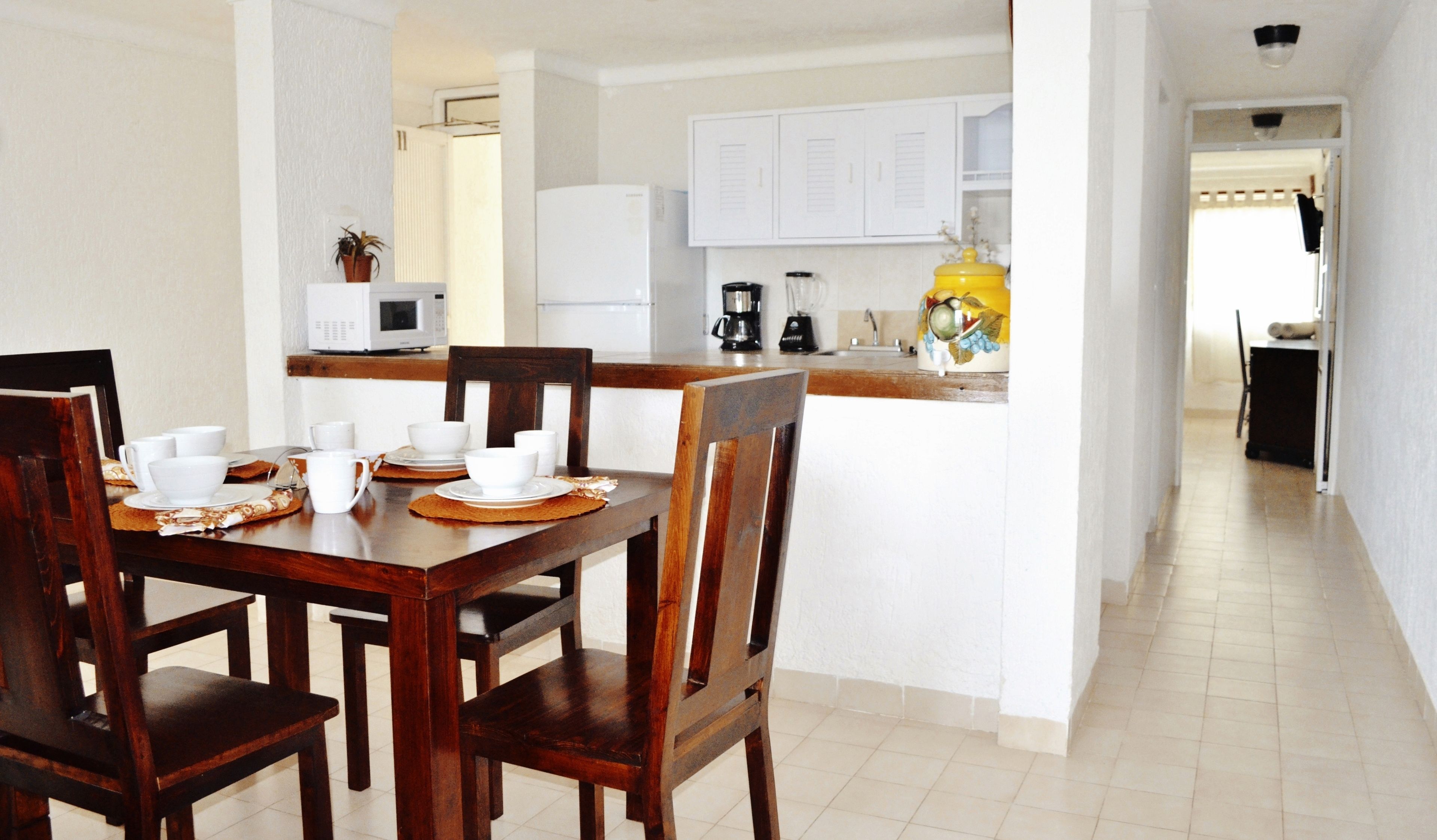 A dining area with a wooden table and chairs, a kitchen in the background, and a hallway leading to another room.
