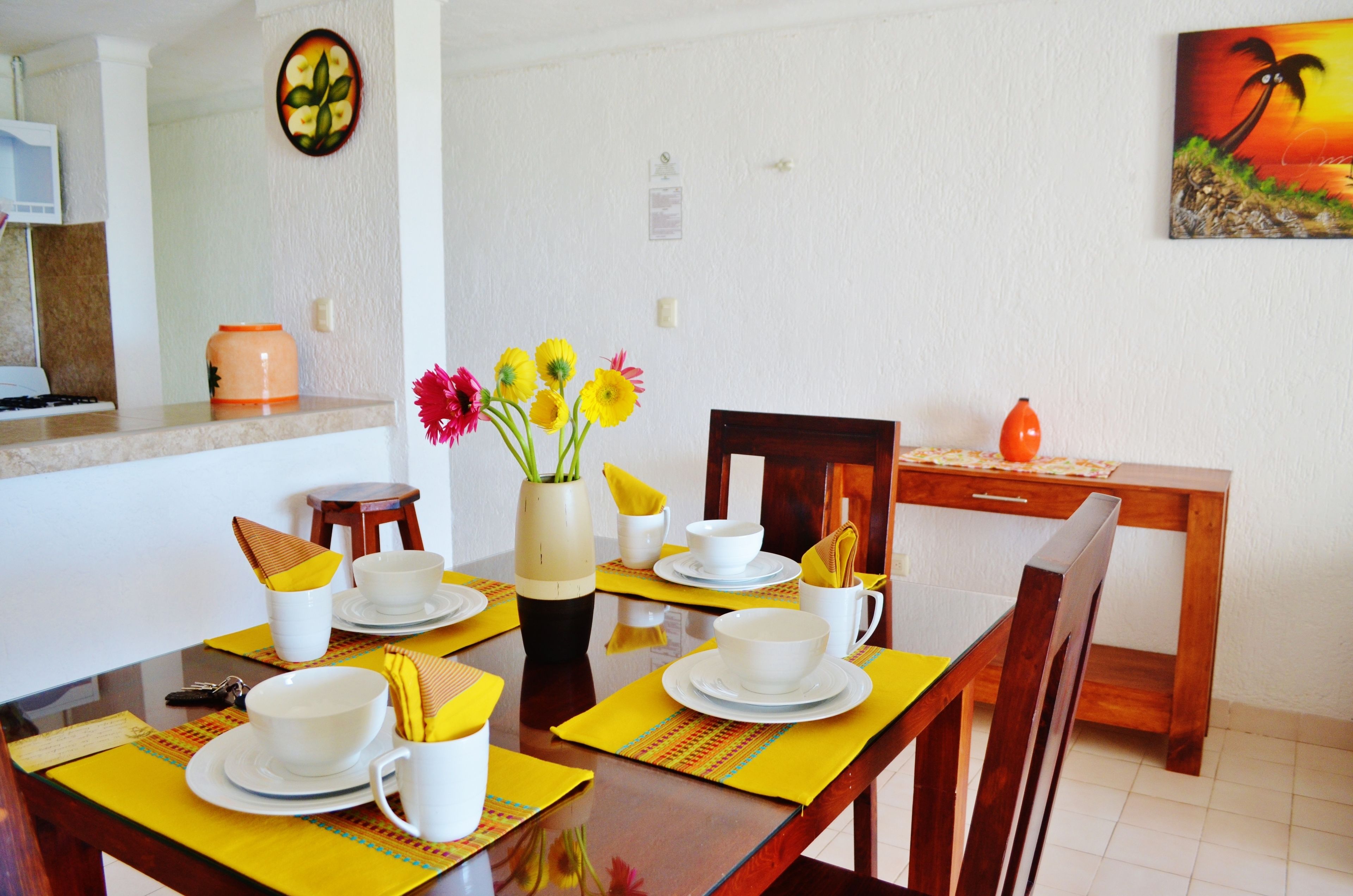 A dining area with a table set for four, including plates, bowls, and cups. A vase with flowers is on the table, and there is a wooden chair and a small table with a vase on it.