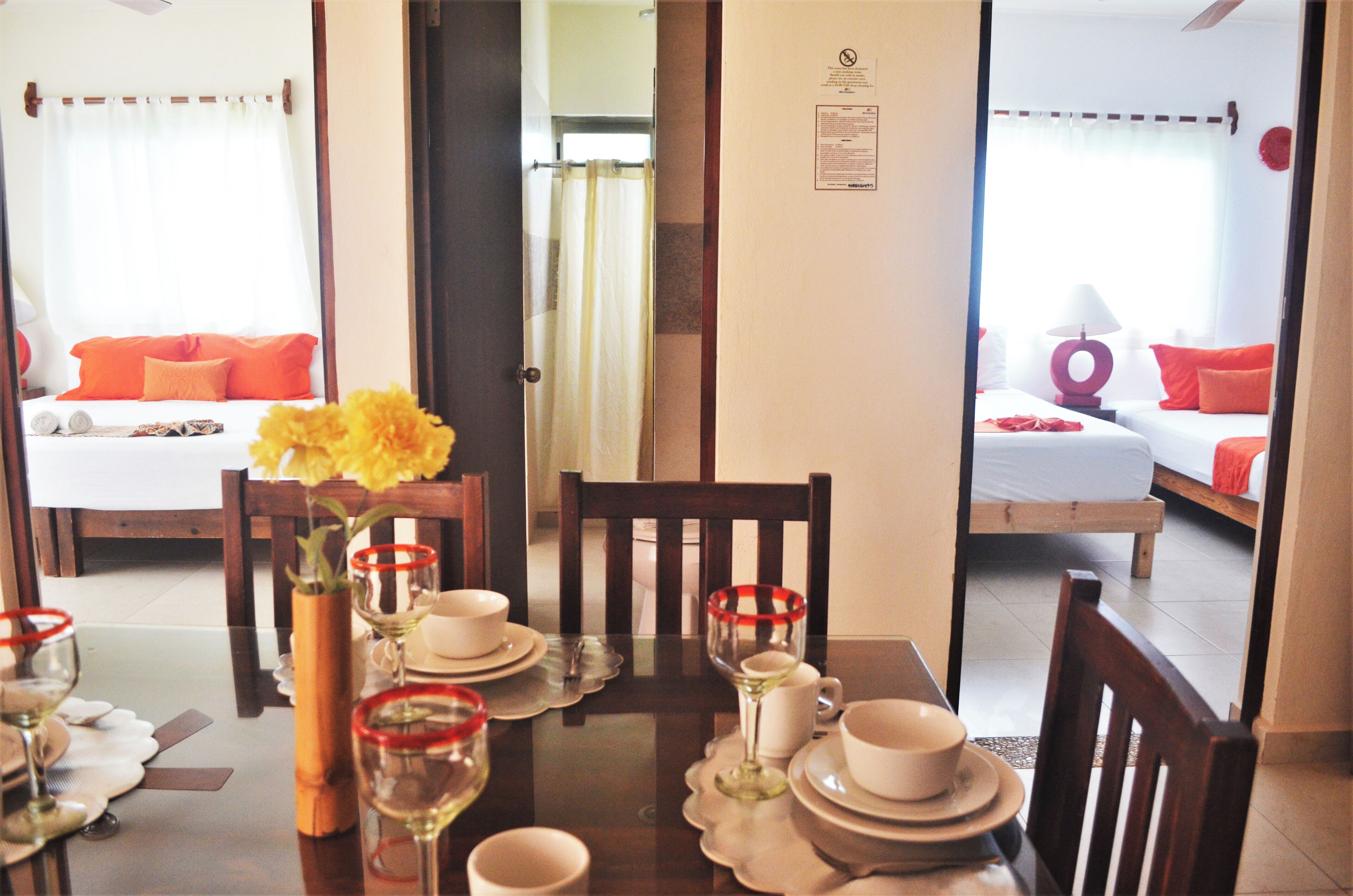 Un comedor con una mesa dispuesta para dos, un florero con flores amarillas, y vista a un dormitorio con una cama y almohadas naranjas.