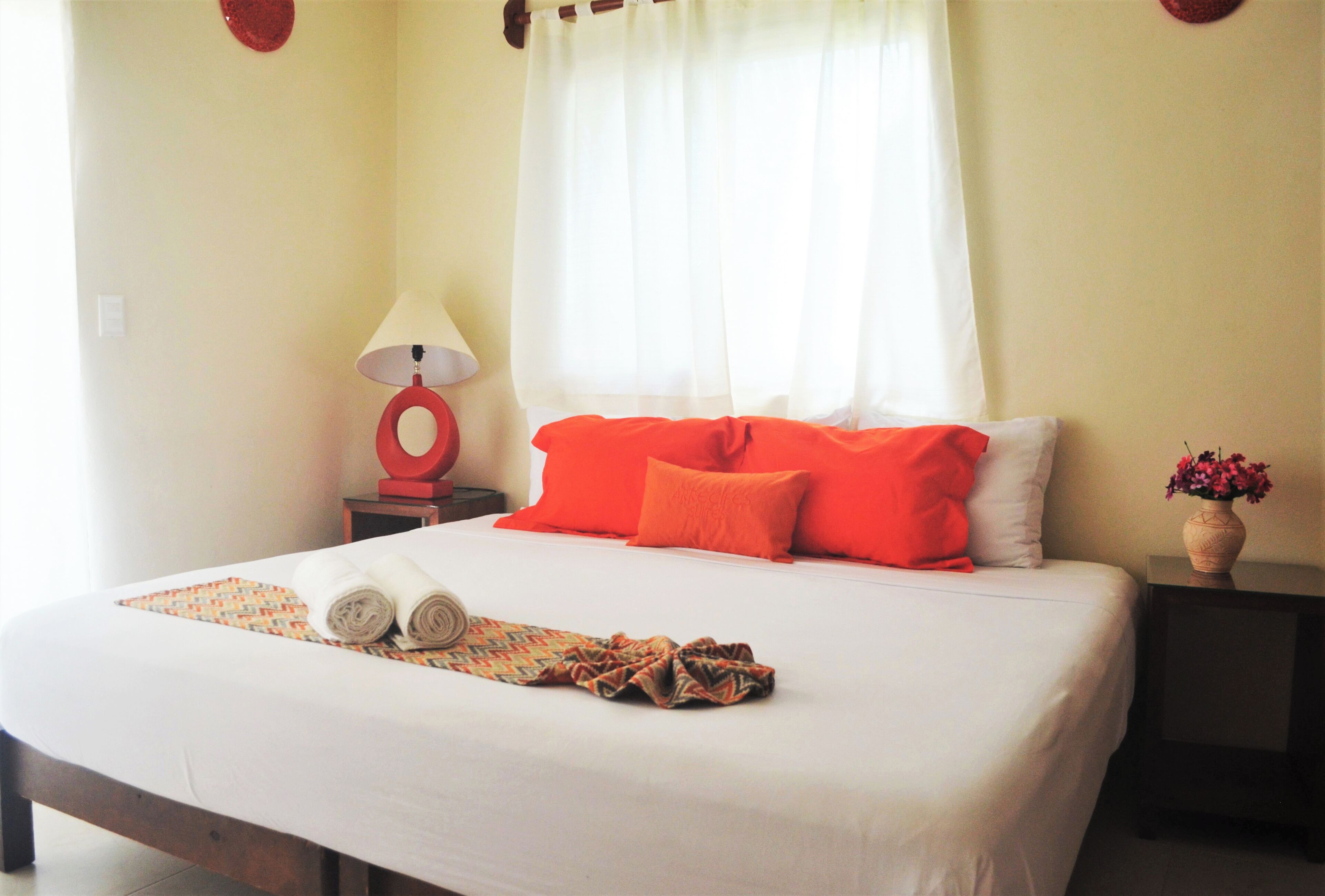 A neatly made bed with rolled towels, a patterned blanket, and bright orange pillows.