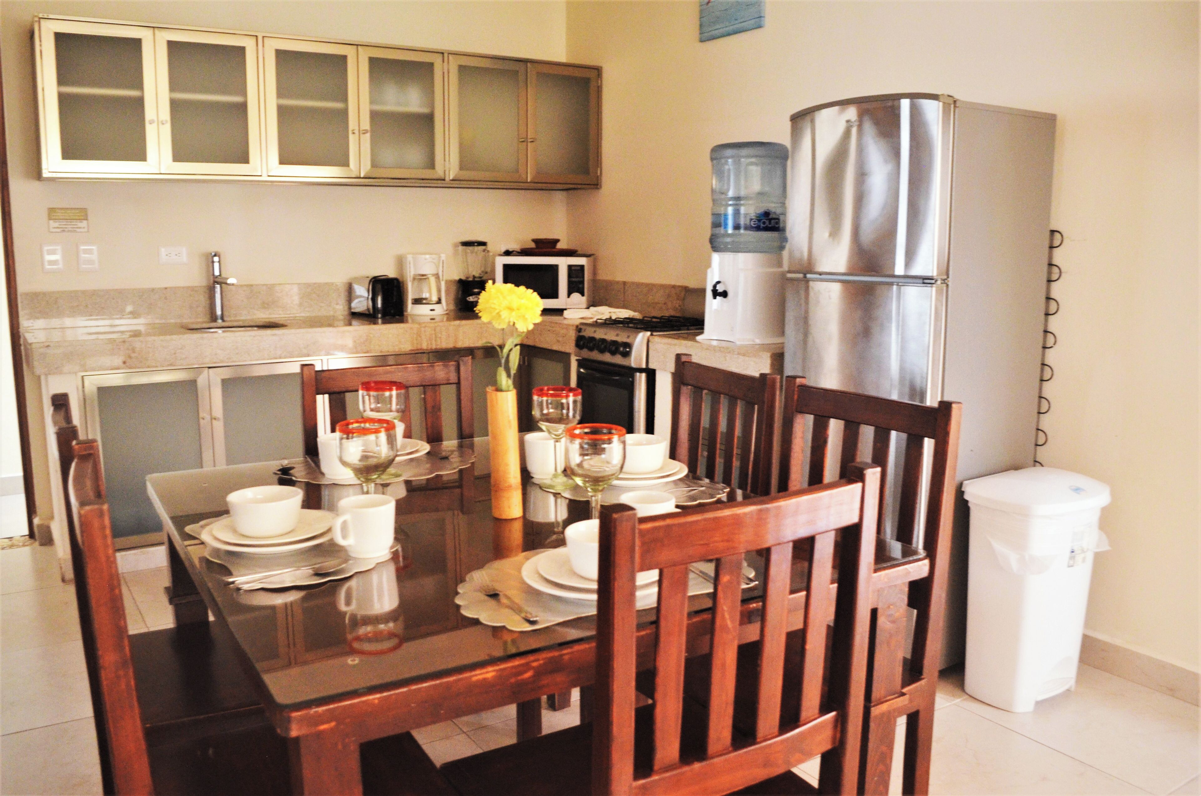 Un comedor con una mesa de madera dispuesta para cuatro personas, un dispensador de agua y un refrigerador.