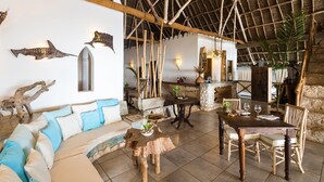 Een ruime kamer met een hoog rieten plafond, een houten eettafel met stoelen, een bank met kussens en decoratieve wandkunst.
