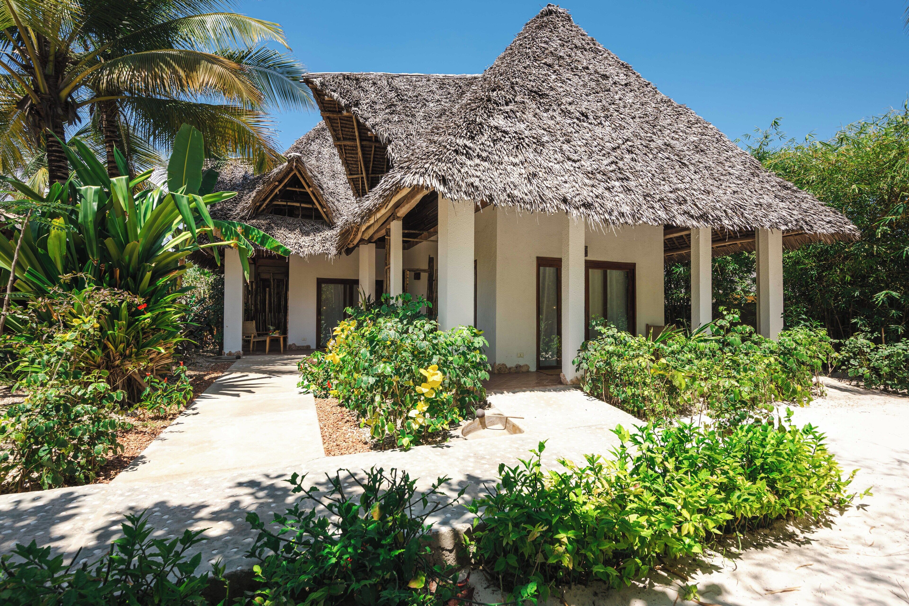 A thatched-roof bungalow with a paved pathway leading to the entrance, surrounded by lush greenery and palm trees.
