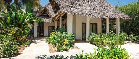 A thatched-roof bungalow with a paved pathway leading to the entrance, surrounded by lush greenery and palm trees.