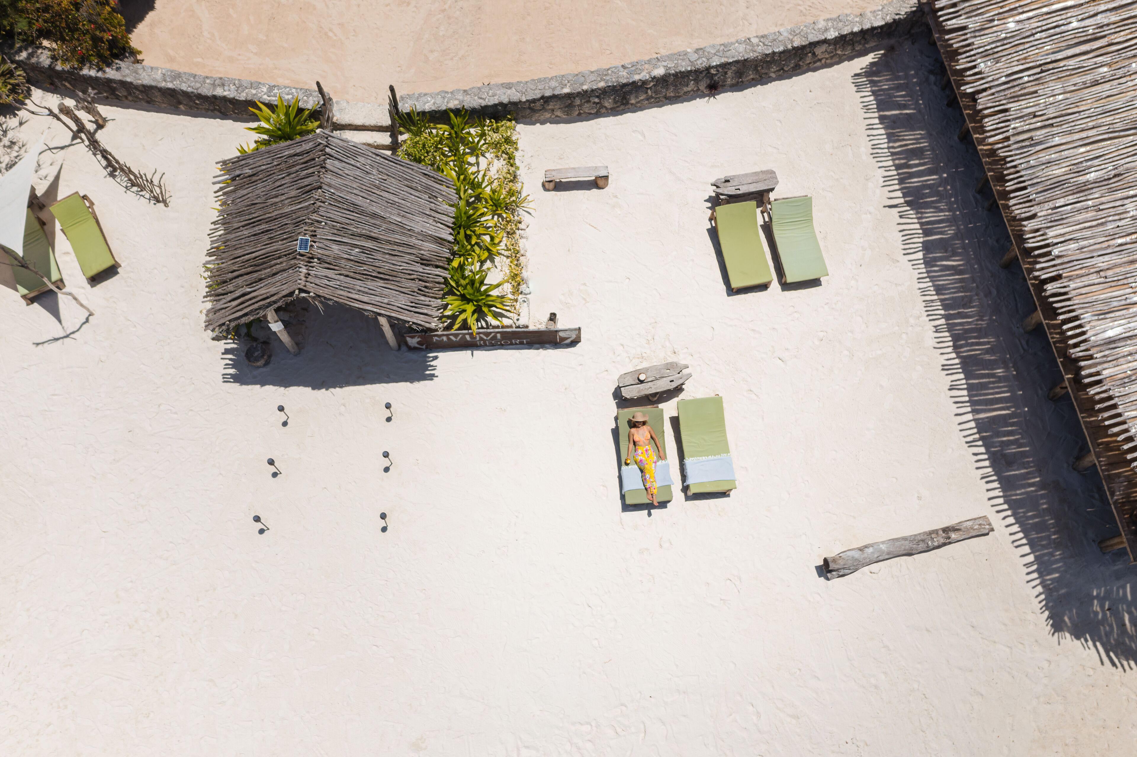 An aerial view of a beach with a thatched-roof hut, lounge chairs, and a small garden area.