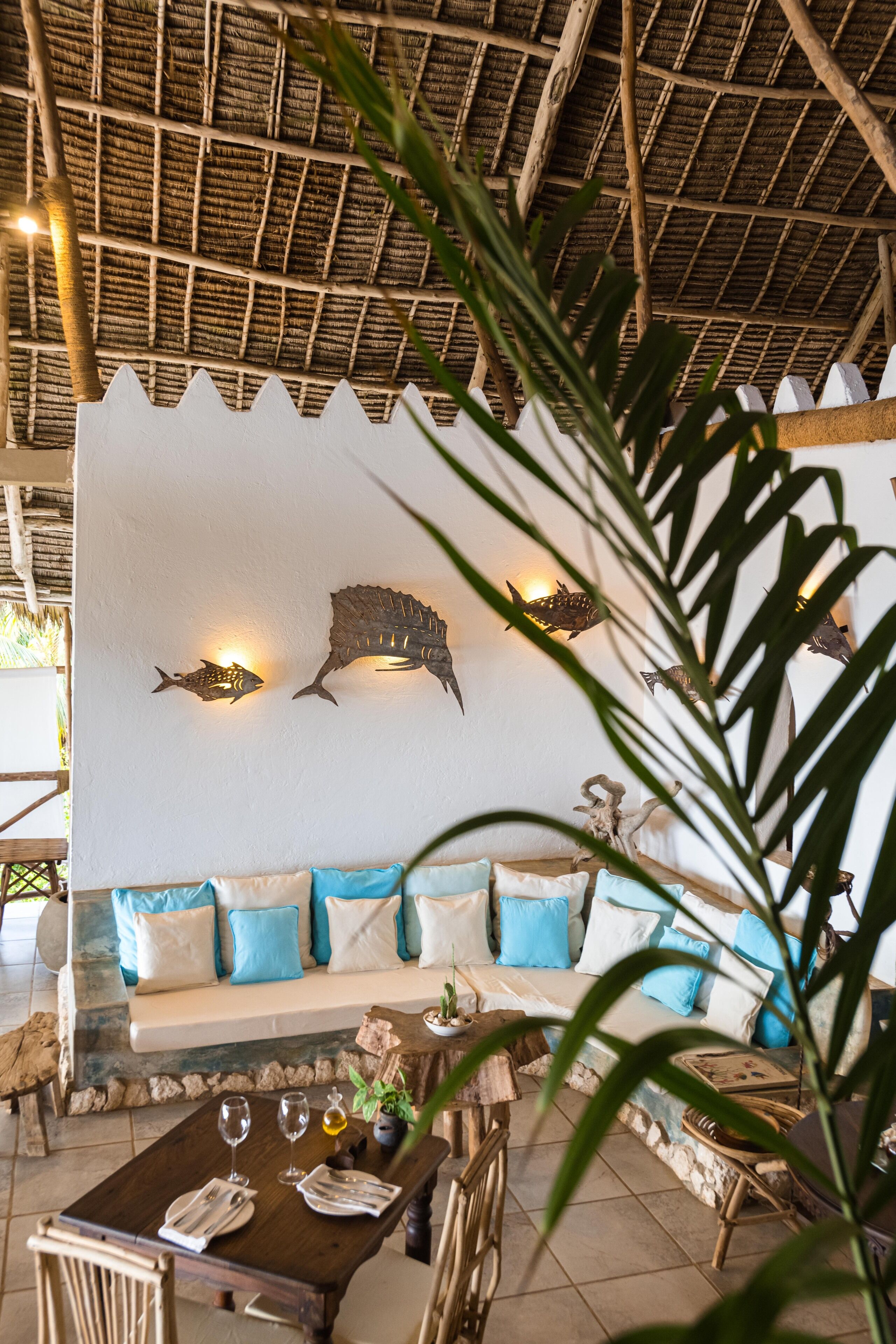 A dining area with a wooden table set for two, a sofa with blue cushions, and a wall with fish-shaped wall art.