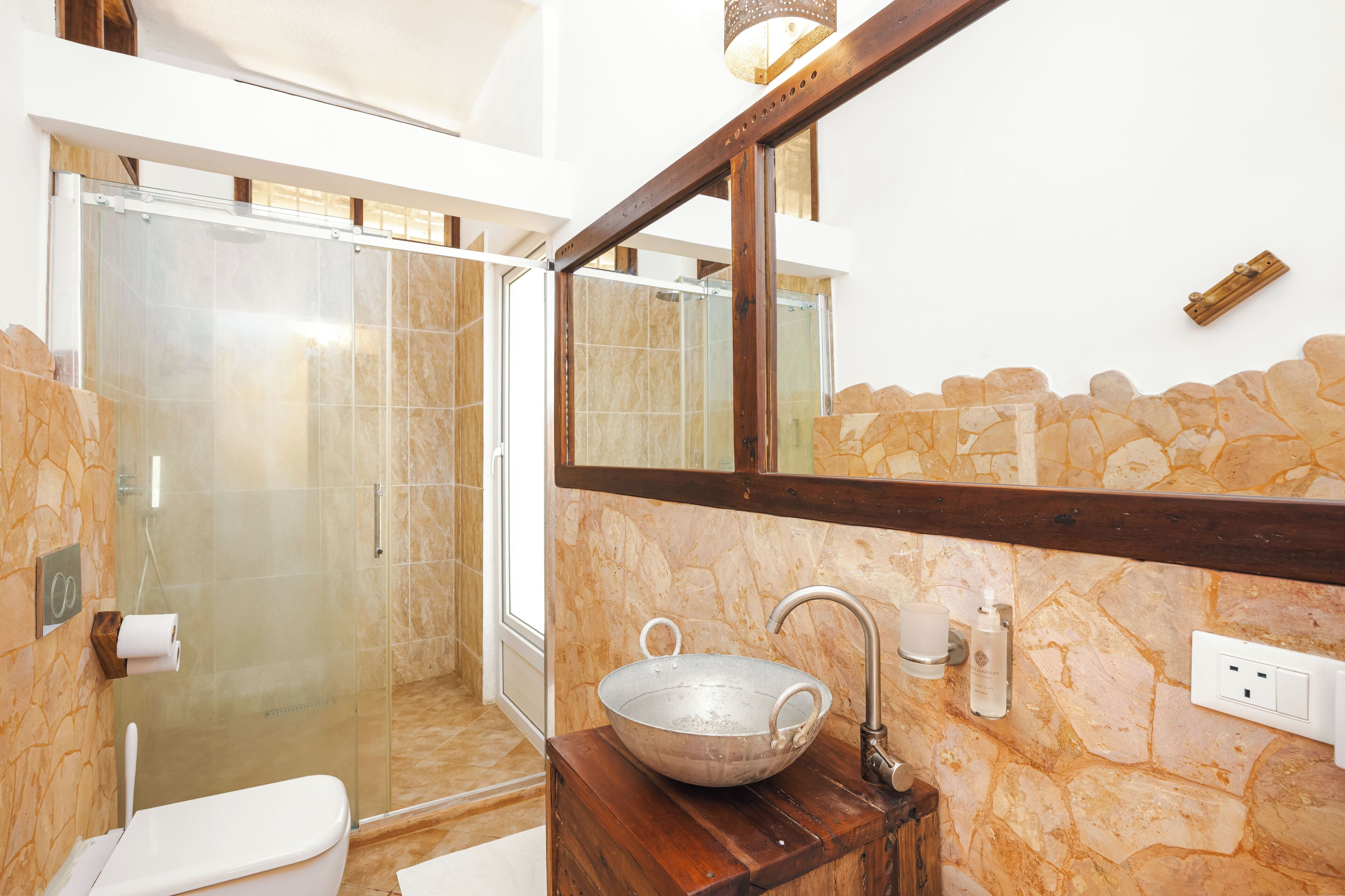 Un baño con pared de piedra, tocador de madera, espejo grande y ducha con mampara de vidrio.