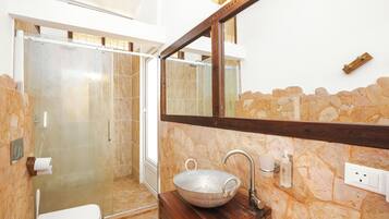 Un baño con pared de piedra, tocador de madera, espejo grande y ducha con mampara de vidrio.