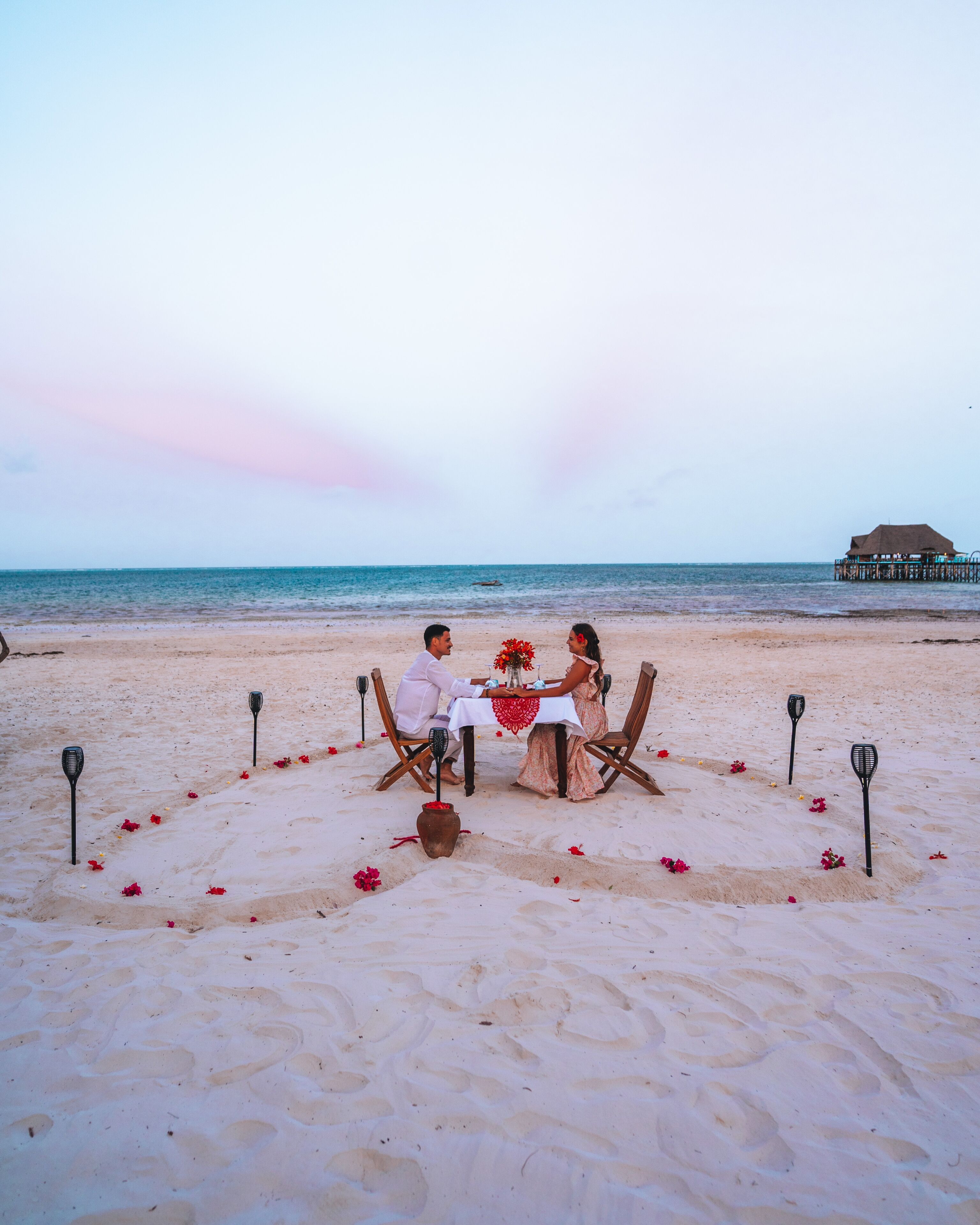 A beachside dining setup with a table and chairs, a vase with flowers, and a decorative fence.