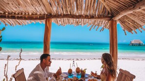 Een houten tafel en stoelen onder een rieten dak, met uitzicht op een strand en helder turquoise water.