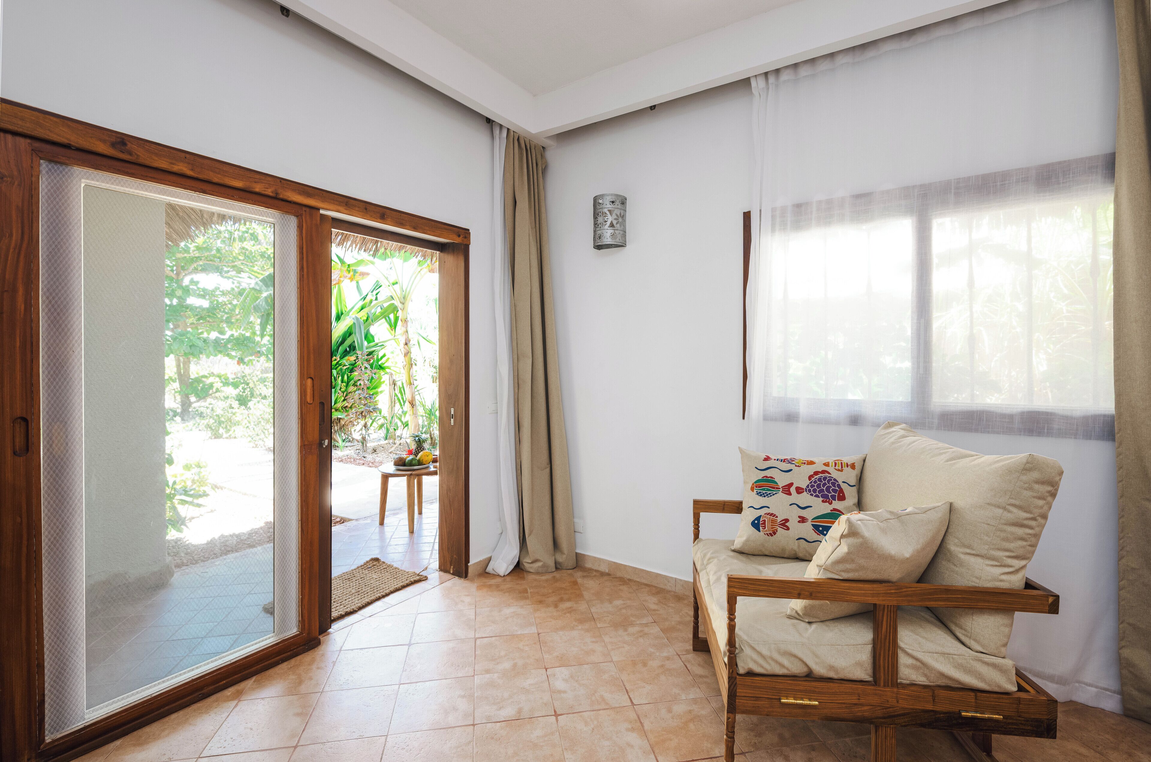 Una habitación con puerta corredera de vidrio que da a un jardín, una silla de madera con cojines y una mesita con una planta.