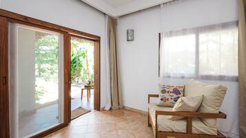 Una habitación con puerta corredera de vidrio que da a un jardín, una silla de madera con cojines y una mesita con una planta.