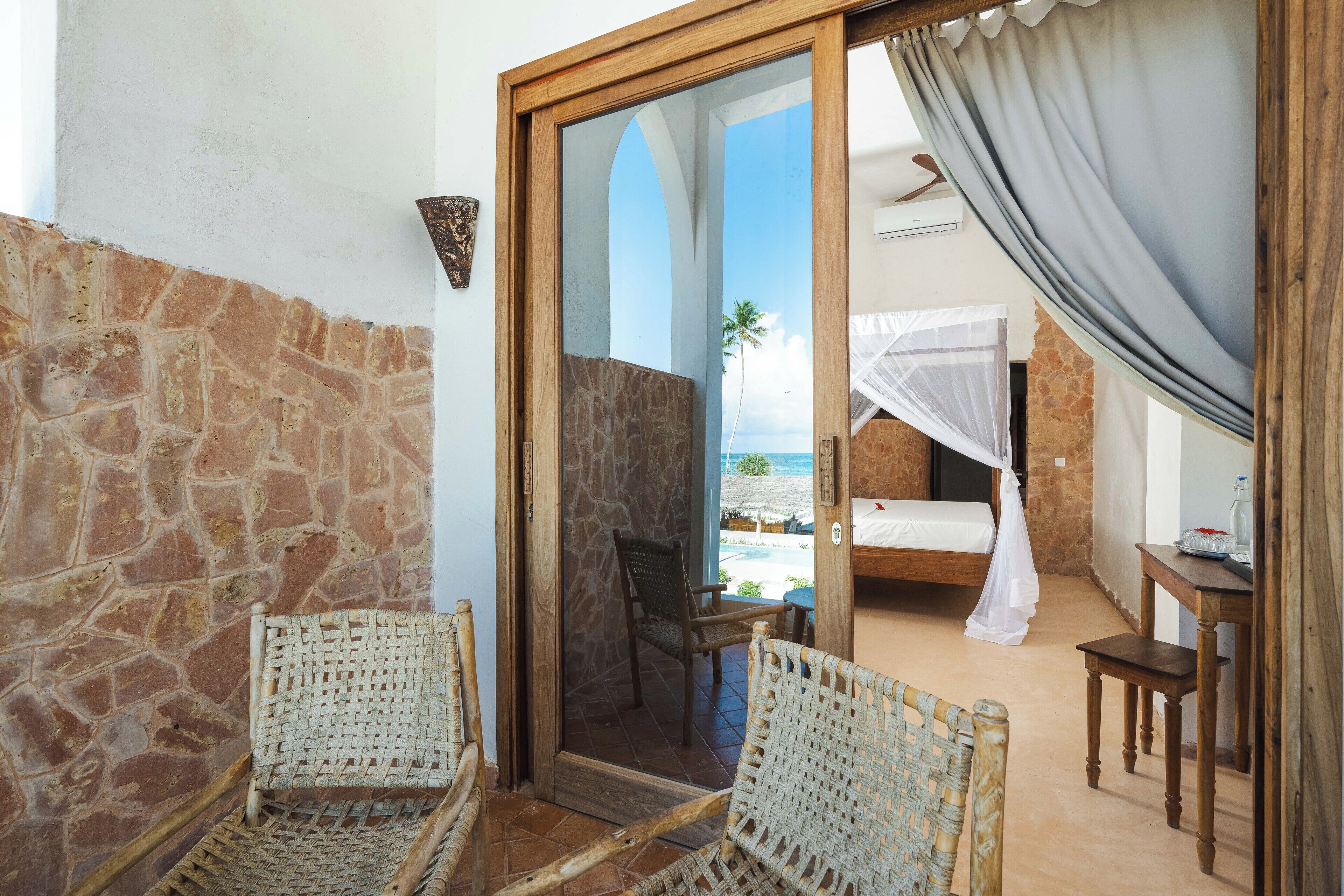 A room with a stone wall, a wooden door leading to a balcony, wicker chairs, a bed with a mosquito net, and a view of the beach.