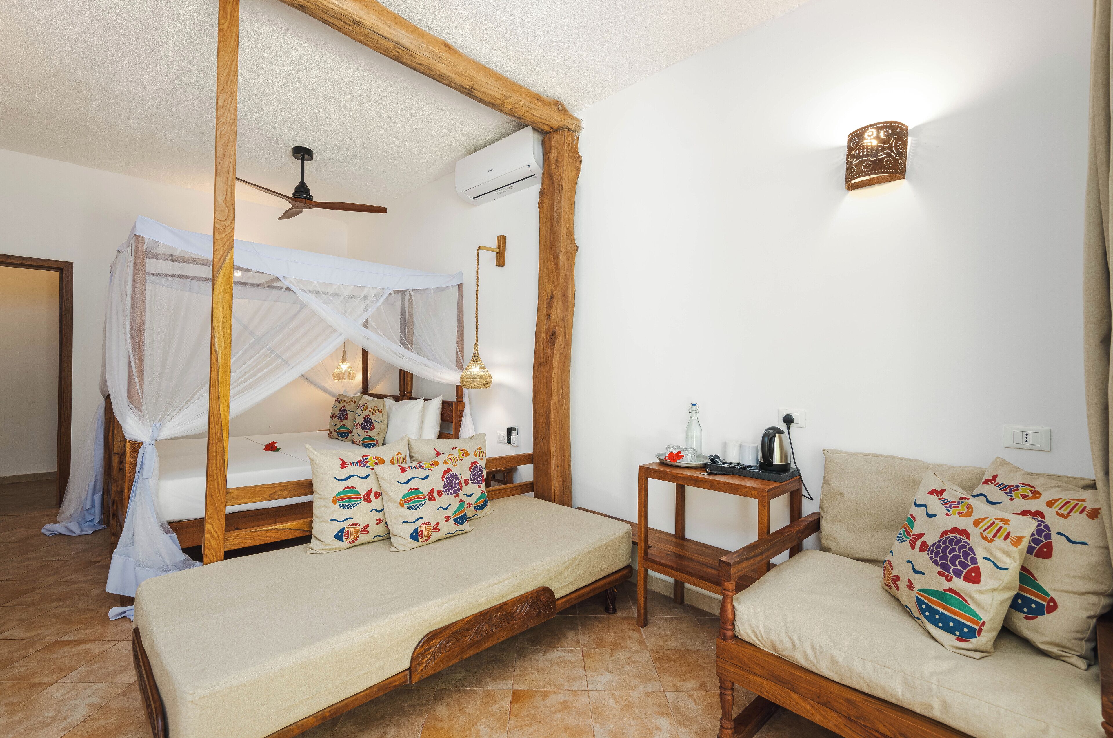 A room with a wooden bed with a canopy, a sofa with colorful pillows, a small wooden table, and a ceiling fan.