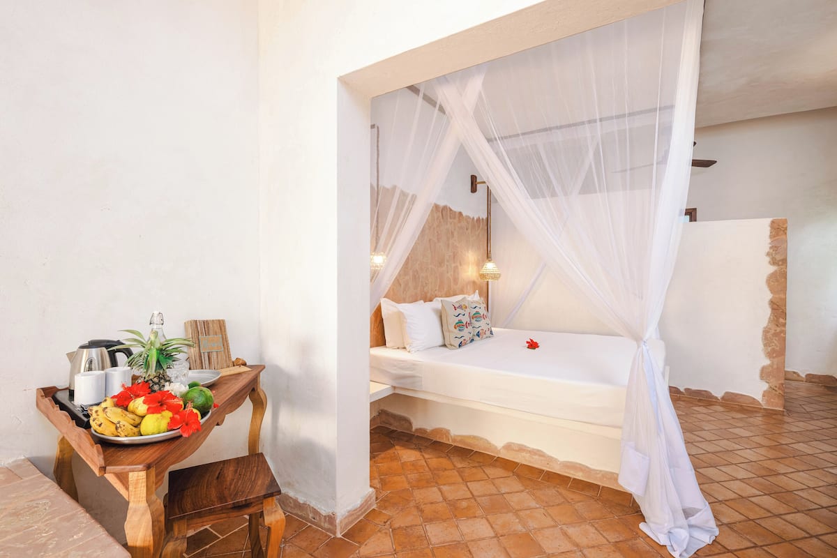 Une chambre à coucher avec un lit sous un dais, une table en bois sur laquelle sont disposés des fruits et une bouilloire, et un sol carrelé.