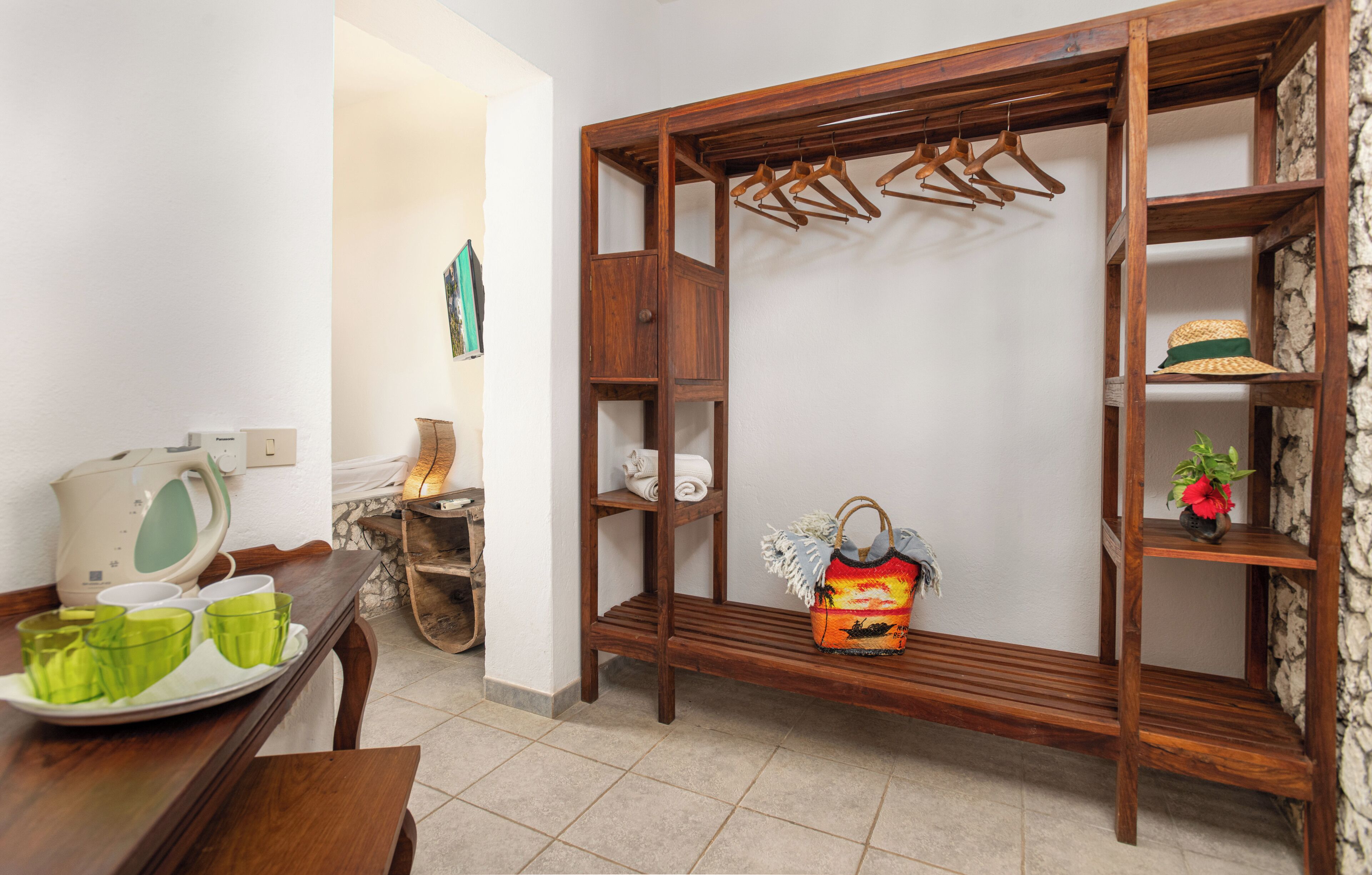 A room with a wooden wardrobe, a bench, a tray with green cups, and a hanging rack with clothes.